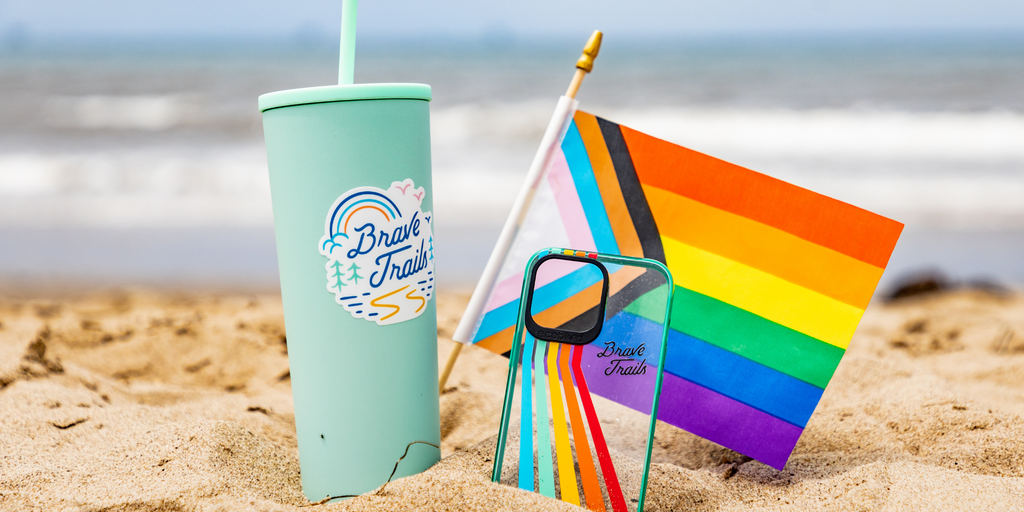 Pride Flag, Reusable Cup With Brave Trails Sticker & Phone Case With Trailblazer Design From EcoBlvd X Brave Trails Collab On a Sandy Beach