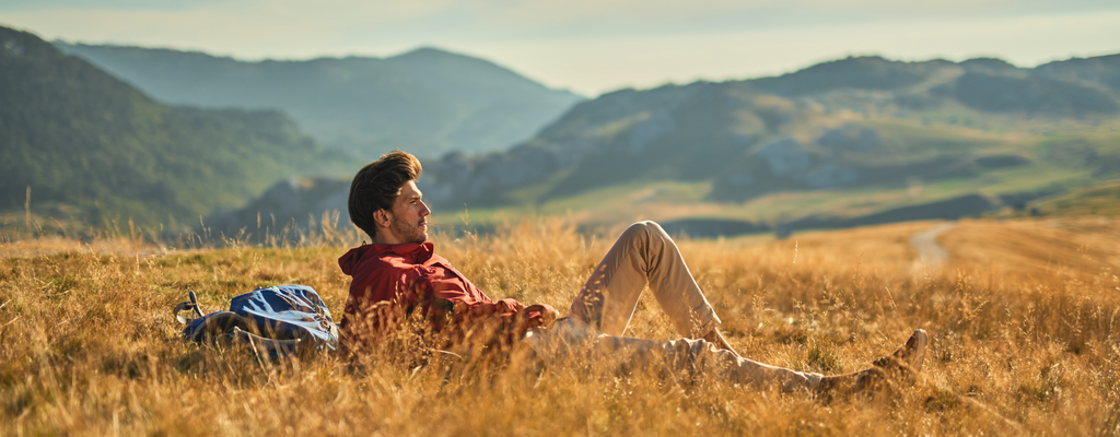 Man in red shirt and khaki pants sitting in golden wheat field with mountain backdrop on a sunny day